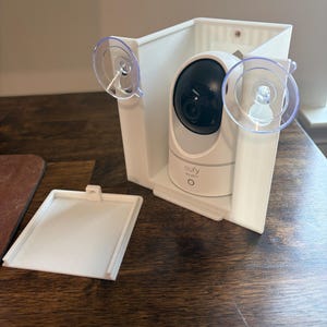 May include: A white security camera with a black lens is mounted inside a white bracket with two clear suction cups. The bracket is on a wooden surface. A white square cover is next to the bracket.