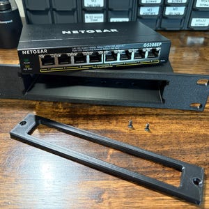 May include: A black Netgear GS308EP network switch with multiple ports, a black rack mount, and a black faceplate. Two small screws are visible. The items are on a wooden surface.