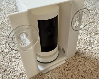 Puede incluir: Soporte blanco para cámara de seguridad con una cámara cilíndrica en blanco y negro en su interior. El soporte tiene dos ventosas transparentes. Fabricado en plástico blanco.