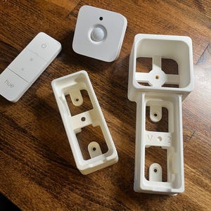 May include: A set of white plastic parts for a smart home device. The set includes a motion sensor, a remote control, and two wall plates. The remote control has the word "hue" printed on it.