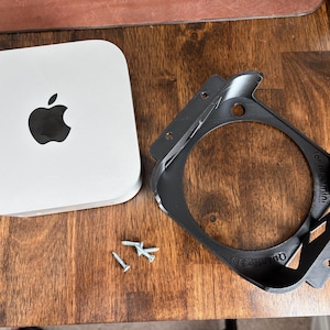 May include: A white Apple Mac Mini computer with the Apple logo, a black bracket, and two screws on a wooden surface. The bracket has the text "for mac mini!" and "Quanta3D" printed on it.