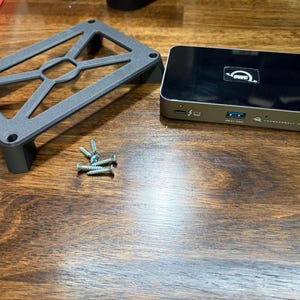 May include: A black and silver Thunderbolt hub with the OWC logo, a gray metal stand, and a small pile of screws. The hub has multiple ports, including USB and Thunderbolt. The stand is rectangular with a geometric design.