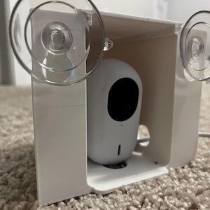May include: A white security camera with a large black lens is housed in a white, rectangular enclosure. Two clear suction cups are attached to the enclosure. A white cable extends from the bottom of the camera.