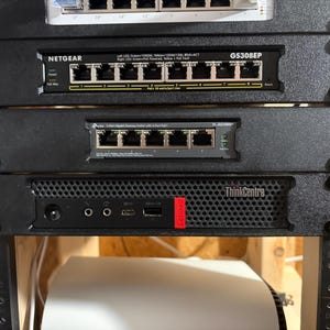May include: A network rack with multiple components, including a Netgear GS308EP switch, a TP-Link switch, and a ThinkCentre computer. The rack is mounted on a wooden frame. The ThinkCentre has a red Lenovo label.