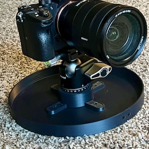 May include: A black Sony digital camera with a zoom lens is mounted on a black tripod head and a circular base. The lens has the text "FE 4/24-105 G OSS". The camera is on a carpeted surface.