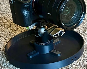 May include: A black Sony digital camera with a zoom lens is mounted on a black tripod head and a circular base. The lens has the text "FE 4/24-105 G OSS". The camera is on a carpeted surface.