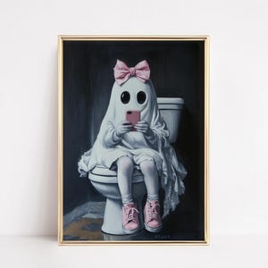 May include: A framed art print depicts a ghost wearing a pink bow and sneakers, sitting on a toilet while looking at a pink smartphone. The ghost is draped in a white sheet. The background is dark with a gold frame.