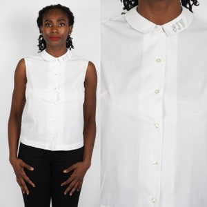 May include: White sleeveless blouse with a rounded collar and button-down front. The collar features embroidered lettering. The blouse is paired with black pants. The image shows a full-length view and a close-up of the blouse.