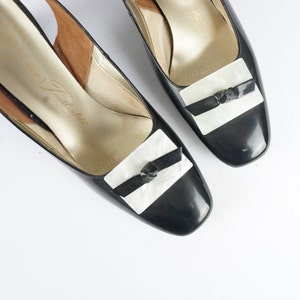 Puede incluir: Par de zapatos de charol negro con correa en el tobillo y detalles de lazo blanco y negro. Los zapatos tienen un tacón bajo y una punta cuadrada.