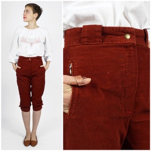 May include: A pair of high-waisted, rust-colored corduroy pants with a zippered pocket on the left side. The pants are cropped above the knee and have a wide leg.