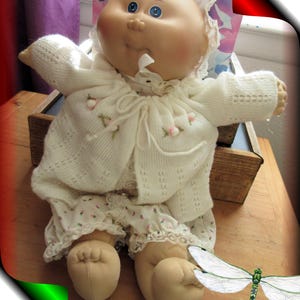 May include: A vintage doll with a round face, blue eyes, and a soft expression. The doll is dressed in a white knitted sweater, matching bonnet, and floral patterned bloomers. The doll is sitting on a wooden surface.