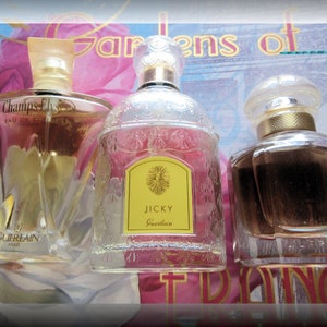May include: Three glass perfume bottles with gold accents. The bottles are filled with clear liquid. The bottles are labeled "Champs Elysees Eau de Toilette Guerlain Paris", "Jicky Guerlain", and "Guerlain".