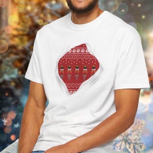 Può includere: T-shirt bianco con un design unico. Un motivo a maglia rosso è visibile attraverso uno strappo, con schiaccianoci e motivi a fiocco di neve. Il design evoca un tema festivo natalizio.