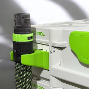 Op de afbeelding: Een groene en zwarte gereedschaphouder met een zwart en groen gereedschap dat eraan is bevestigd. De gereedschaphouder is bevestigd aan een grijze en groene gereedschapskist met het woord "Festool" erop gedrukt.