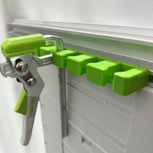 Può includere: Un morsetto Festool verde lime e argento attaccato a un supporto verde. Il supporto è montato su un sistema a binario bianco e argento. Sul morsetto è stampata la parola "Festool".
