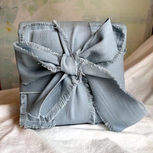 Puede incluir: Una caja de regalo de tela azul claro con un borde deshilachado y un gran lazo atado en la parte superior.