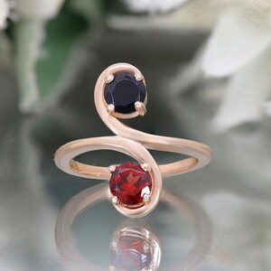 May include: A rose gold ring with a black and red gemstone design. The ring has a unique, curved shape with a black gemstone on one side and a red gemstone on the other.