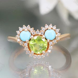 Op de afbeelding: Een gouden ring met een hartvormig ontwerp met een grote groene edelsteen omringd door kleinere witte edelstenen. Twee kleinere turquoise edelstenen zijn aan weerszijden van de groene edelsteen geplaatst.