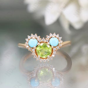 Op de afbeelding: Een gouden ring met een hartvormig ontwerp met een groene peridoot edelsteen omringd door kleinere diamanten en twee turquoise edelstenen.