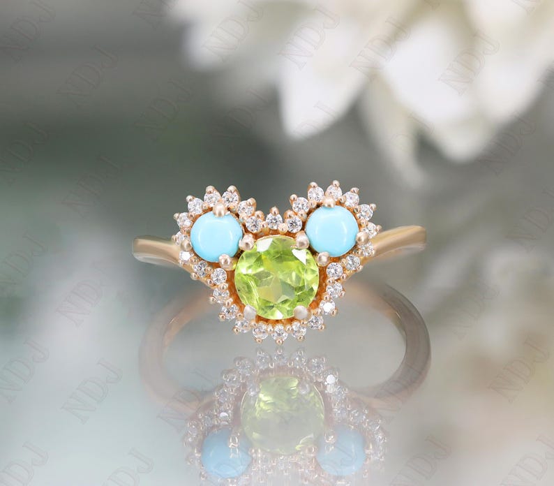 Op de afbeelding: Een gouden ring met een hartvormig ontwerp met een groene peridoot edelsteen omringd door kleinere diamanten en twee turquoise edelstenen.