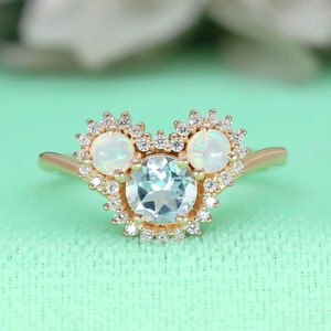 May include: A gold ring with a large aquamarine center stone surrounded by smaller diamonds. The ring has two smaller opal stones on either side of the center stone, also surrounded by diamonds.