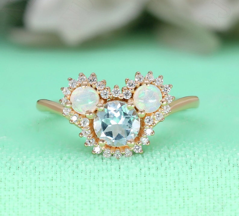 May include: A gold ring with a large aquamarine center stone surrounded by smaller diamonds. The ring has two smaller opal stones on either side of the center stone, also surrounded by diamonds.