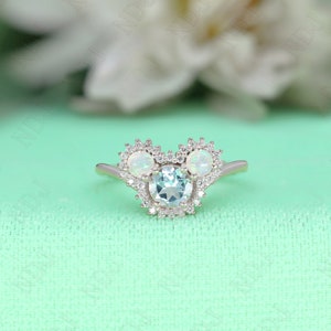 May include: A silver ring with a blue gemstone surrounded by smaller white gemstones. The ring is shaped like a Mickey Mouse head with two smaller gemstones for the ears.