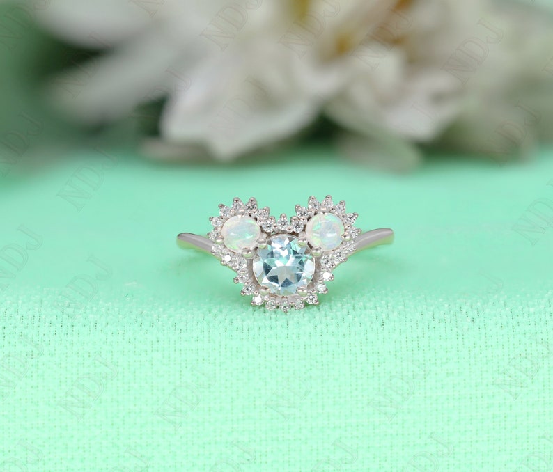 May include: A silver ring with a blue gemstone surrounded by smaller white gemstones. The ring is shaped like a Mickey Mouse head with two smaller gemstones for the ears.