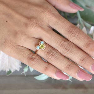 Op de afbeelding: Een gouden ring met een hartvormige zetting met een groene edelsteen omringd door kleinere witte edelstenen en twee turquoise edelstenen.