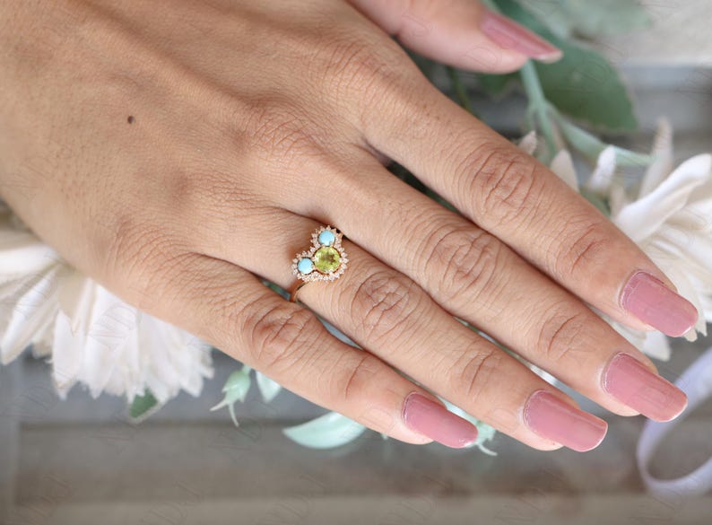 Op de afbeelding: Een gouden ring met een hartvormige zetting met een groene edelsteen omringd door kleinere witte edelstenen en twee turquoise edelstenen.