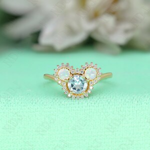 May include: A gold ring with a light blue gemstone surrounded by smaller clear gemstones. Two white opals are set on either side of the center stone, each surrounded by smaller clear gemstones.