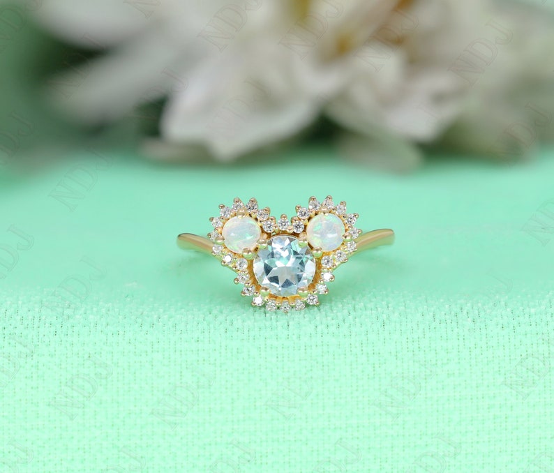May include: A gold ring with a light blue gemstone surrounded by smaller clear gemstones. Two white opals are set on either side of the center stone, each surrounded by smaller clear gemstones.