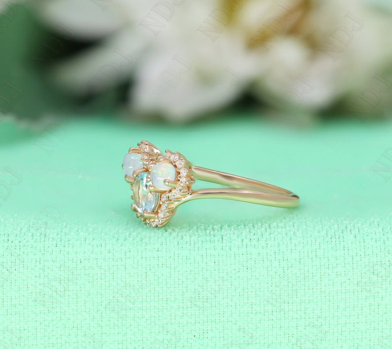 May include: A delicate gold ring with three oval-shaped gemstones. The center stone is a light blue aquamarine, flanked by two white opals. The ring is set with small diamonds around the stones.