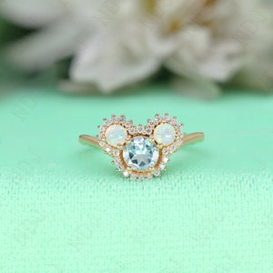 May include: A gold ring with a round aquamarine center stone surrounded by a halo of small diamonds. The ring has two smaller round opal stones on either side of the center stone, also surrounded by halos of small diamonds.