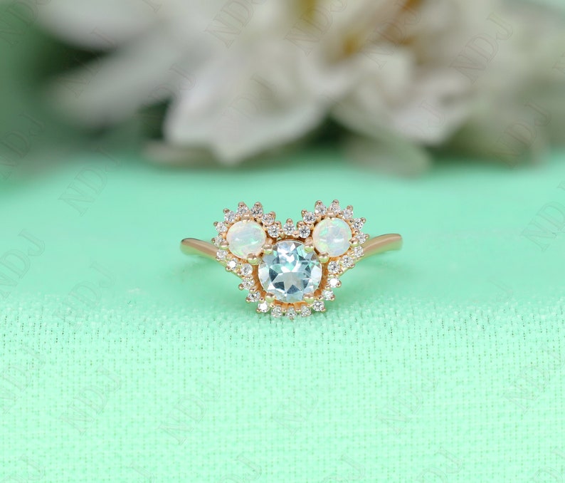 May include: A gold ring with a round aquamarine center stone surrounded by a halo of small diamonds. The ring has two smaller round opal stones on either side of the center stone, also surrounded by halos of small diamonds.