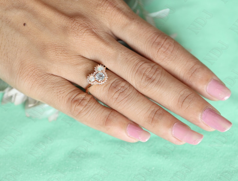 May include: A heart-shaped ring with a large, clear center stone surrounded by smaller, clear stones. The ring is set in rose gold and is worn on a hand with pink nail polish.