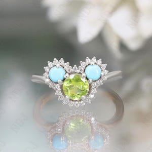 Op de afbeelding: Een zilveren ring met een groene peridoot-edelsteen omringd door kleine diamanten. De ring heeft ook twee turquoise edelstenen aan weerszijden van de peridoot.