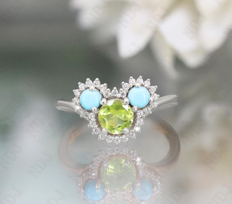 Op de afbeelding: Een zilveren ring met een groene peridoot-edelsteen omringd door kleine diamanten. De ring heeft ook twee turquoise edelstenen aan weerszijden van de peridoot.