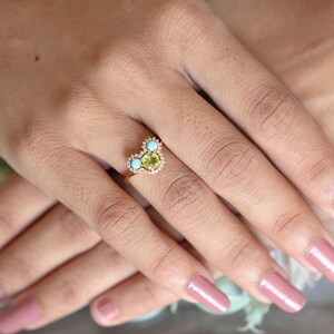 Op de afbeelding: Een gouden ring met een hartvormige zetting met een groene edelsteen omringd door kleinere diamanten en twee turquoise edelstenen.