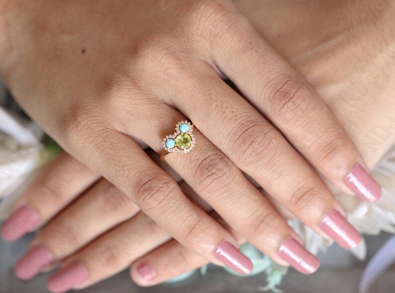 Op de afbeelding: Een gouden ring met een hartvormige zetting met een groene edelsteen omringd door kleinere diamanten en twee turquoise edelstenen.