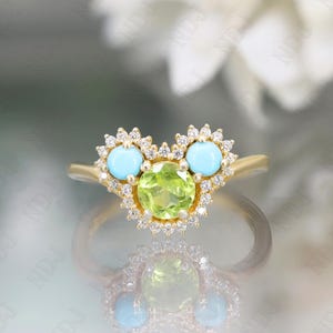 Op de afbeelding: Een gouden ring met een groene peridoot edelsteen omringd door kleine diamanten. De ring heeft ook twee turquoise edelstenen, elk omringd door kleine diamanten.