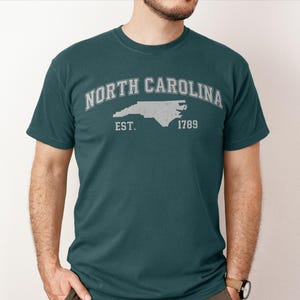 May include: Teal t-shirt with a distressed white graphic. The graphic features the words "NORTH CAROLINA" arched above a state outline, with "EST. 1789" below. The shirt is a crew neck and has short sleeves.