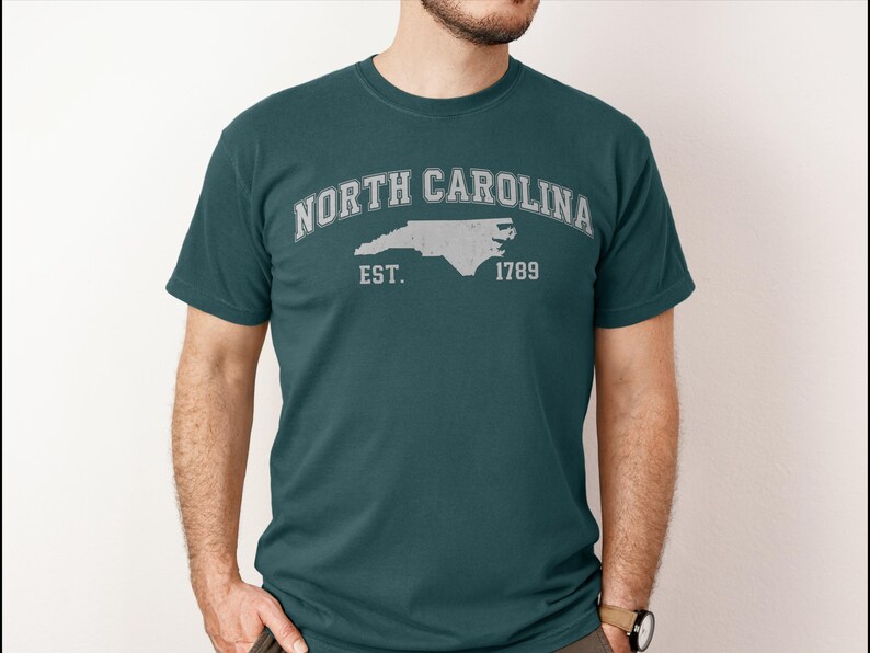 May include: Teal t-shirt with a distressed white graphic. The graphic features the words "NORTH CAROLINA" arched above a state outline, with "EST. 1789" below. The shirt is a crew neck and has short sleeves.