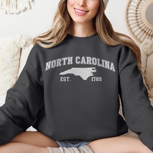 May include: Dark gray sweatshirt with the text "NORTH CAROLINA" in arched white letters above a white outline of the state, with "EST. 1789" below. The sweatshirt is a casual style.