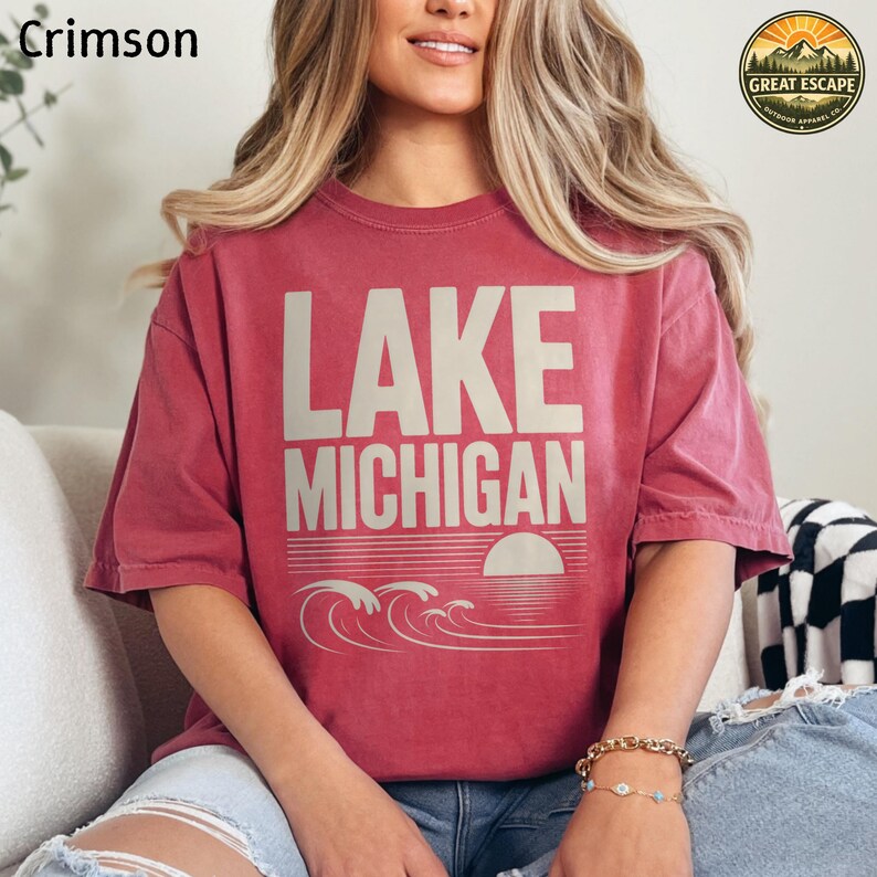 K&ouml;nnte beinhalten: Ein kurz&auml;rmeliges T-Shirt in Karmesinrot mit dem Text LAKE MICHIGAN in gro&szlig;en beigefarbenen Buchstaben. Unter dem Text befindet sich eine Grafik einer untergehenden Sonne &uuml;ber stilisierten Wellen. Das T-Shirt hat einen Rundhalsausschnitt und eine lockere Passform.