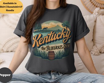 May include: Dark gray t-shirt featuring a Kentucky design with a landscape scene, the text "Kentucky the Bluegrass State", and a barrel. Available in T-Shirt, Sweatshirt & Hoodie.