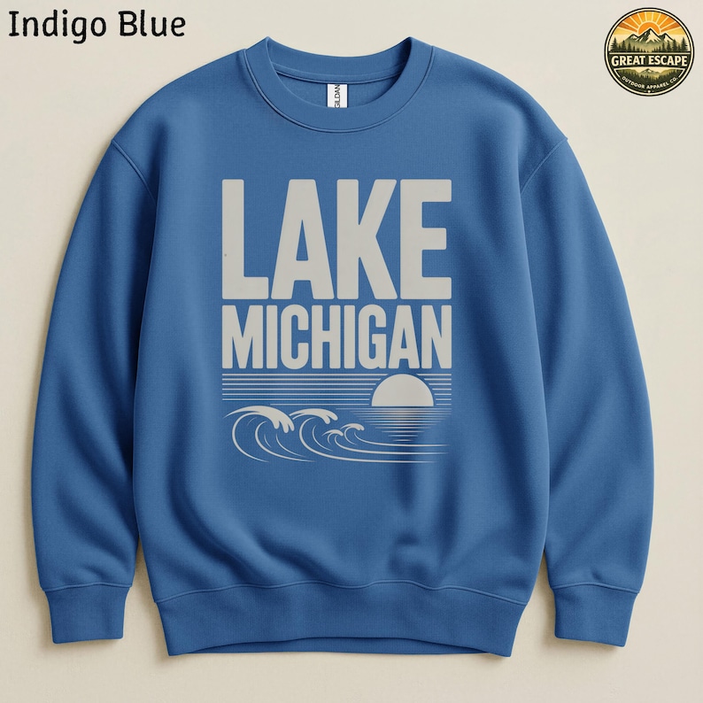 K&ouml;nnte beinhalten: Ein indigoblauer Rundhals-Pullover mit dem wei&szlig;en Schriftzug LAKE MICHIGAN &uuml;ber einer Grafik aus stilisierten Wellen und einer untergehenden Sonne. In der oberen rechten Ecke befindet sich ein kreisf&ouml;rmiges Logo mit dem Text GREAT ESCAPE OUTDOOR APPAREL CO.