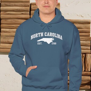 May include: A teal hoodie with a front pocket and a drawstring hood. The front of the hoodie features the words "North Carolina" in a curved font, with a white outline of the state and the text "Est. 1789" below it.