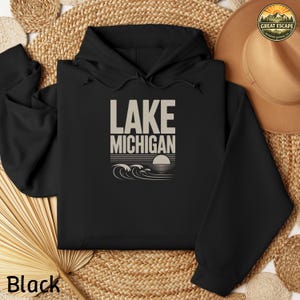 K&ouml;nnte beinhalten: Ein schwarzer Kapuzenpullover mit einem beigen Grafikdruck auf der Brust. Der Text lautet LAKE MICHIGAN &uuml;ber einer stilisierten Illustration einer untergehenden Sonne und Meereswellen. Der Hoodie hat eine Kapuze mit Kordelzug und lange &Auml;rmel.