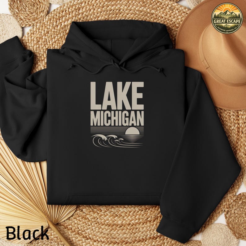 K&ouml;nnte beinhalten: Ein schwarzer Kapuzenpullover mit einem beigen Grafikdruck auf der Brust. Der Text lautet LAKE MICHIGAN &uuml;ber einer stilisierten Illustration einer untergehenden Sonne und Meereswellen. Der Hoodie hat eine Kapuze mit Kordelzug und lange &Auml;rmel.